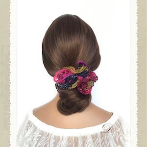 May include: A crocheted hair scrunchie in shades of pink, purple, green, and black. The scrunchie is tied around a woman's brown hair.