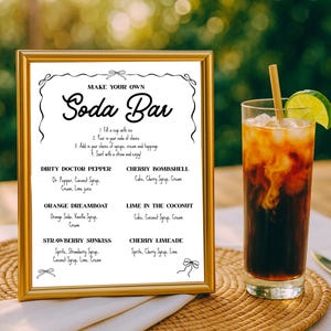 May include: A gold-framed sign with the words "MAKE YOUR OWN Soda Bar" and a list of soda recipes. A tall glass of soda with ice, a lime wedge, and a straw sits next to the sign. The sign is on a woven mat.