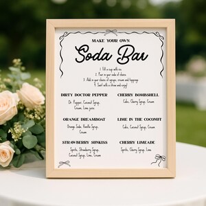 May include: A framed sign with the words "MAKE YOUR OWN Soda Bar" in elegant script. The sign lists soda bar recipes like "Dirty Doctor Pepper" and "Cherry Bombshell" with ingredients. The frame is light wood, and the background is blurred.