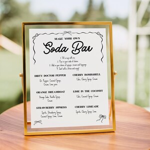 May include: A gold-framed sign with the words "MAKE YOUR OWN Soda Bar" in elegant script. The sign lists soda recipes like Dirty Doctor Pepper and Cherry Bombshell. The sign is on a wooden surface.