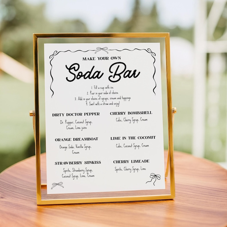 May include: A gold-framed sign with the words "MAKE YOUR OWN Soda Bar" in elegant script. The sign lists soda recipes like Dirty Doctor Pepper and Cherry Bombshell. The sign is on a wooden surface.