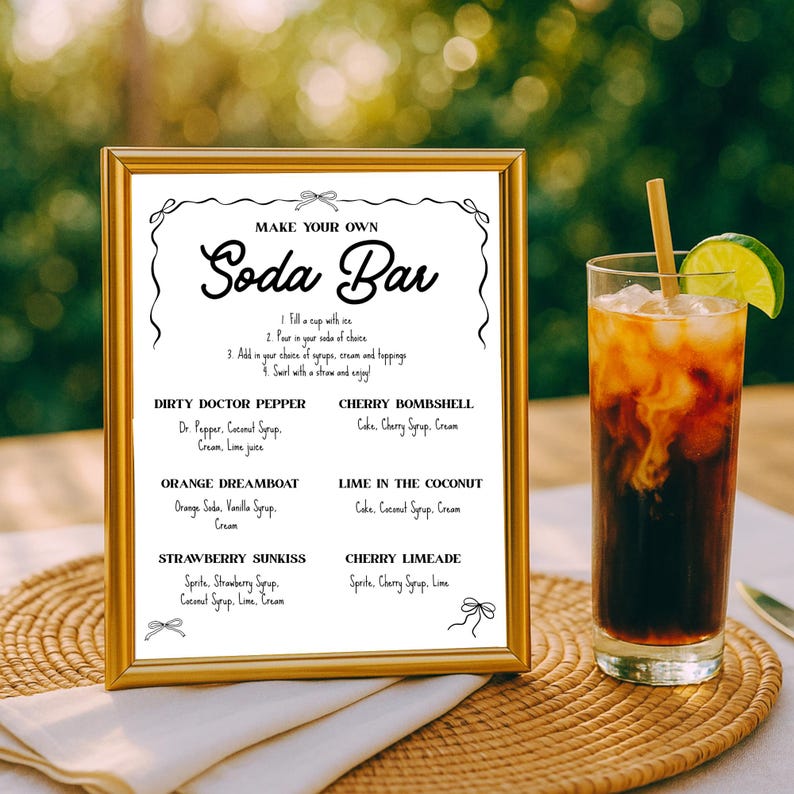 May include: A gold-framed sign with the words "MAKE YOUR OWN Soda Bar" and a list of soda recipes. A tall glass of soda with ice, a lime wedge, and a straw sits next to the sign. The sign is on a woven mat.