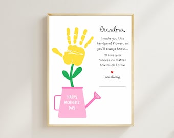 May include: A framed print featuring a handprint flower design. The yellow handprint forms the flower, with a green stem and a pink watering can base. Text includes "Grandma" and "Happy Mother's Day."