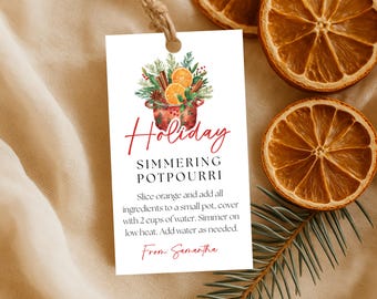 May include: A white tag with the words "Holiday Simmering Potpourri" in red script, featuring a watercolor illustration of a pot with oranges, cinnamon, and greenery. Dried orange slices and a sprig of pine are also visible.