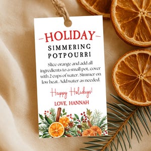 以下が含まれることがあります： 「HOLIDAY SIMMERING POTPOURRI」のテキストと使用方法が記載された白いタグ。タグは紐で結ばれ、オレンジ、シナモンスティック、緑のイラストで装飾されています。乾燥したオレンジのスライスと松の小枝も表示されています。