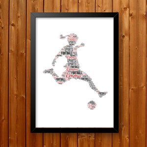May include: Framed artwork featuring a soccer player silhouette composed of the words "Football," "Sports," and the name "Emma" in black and red. The artwork is set against a white background and framed in black, mounted on a wooden wall.