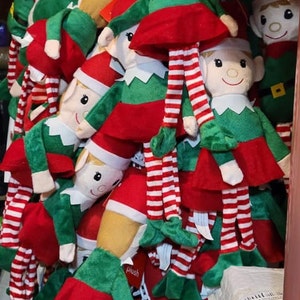 May include: A group of plush elf ornaments with red and green clothing, white and red striped legs, and red hats. The elves are arranged in a pile, with some of them facing forward and others facing sideways.