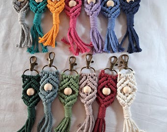 Puede incluir: Una colección de llaveros de macramé en varios colores, incluyendo gris, turquesa, naranja, rosa, morado y azul. Cada llavero presenta una cuenta de madera y una borla, unidos a un cierre de color bronce.