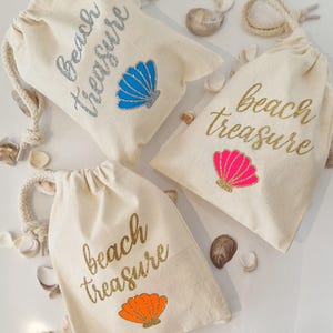 May include: Three beige drawstring bags with the words "beach treasure" in glittery script. Each bag features a seashell graphic in blue, pink, and orange. Seashells are scattered around the bags.