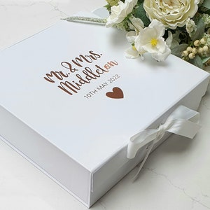May include: A square white gift box featuring a white ribbon closure and copper-coloured text that reads Mr. & Mrs. Middleton 10TH MAY 2022 with a small copper heart icon. The box is decorated with a cluster of white and cream-coloured artificial flowers.