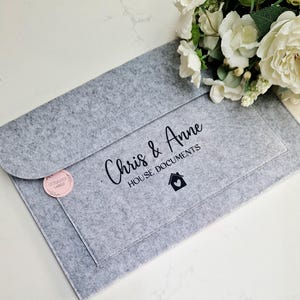 May include: A grey felt folder with the text "Chris & Anne House Documents" and a small house icon with a heart inside. The folder has a pink and white sticker with the text "Little Lucky Labels" on the front.