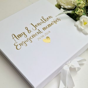May include: White keepsake box with a gold inscription that reads "Amy & Jonathon Engagement memories 29.04.2024" and a gold heart. The box has a white satin ribbon.