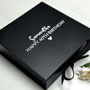 May include: A black gift box with a white ribbon and a white flower. The box has the words "Samantha HAPPY 40TH BIRTHDAY" in white script.