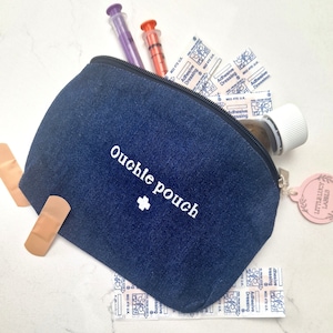 May include: A blue denim pouch with a zipper closure and the text "ouchle pouch" embroidered in white. The pouch is filled with medical supplies, including bandages, gauze, and syringes.