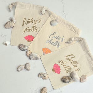 May include: Three off-white cotton drawstring pouches featuring personalised text and shell graphics. The bags are labelled Libby's shells, Evie's shells, and Bella's shells in glittery script, accompanied by pink or orange seashell illustrations.