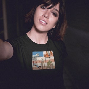 May include: A woman wearing a black t-shirt with a comic book style graphic print of a scene with four panels. The graphic features a person in a white shirt and blue jeans, a person in a red shirt and blue jeans, a person in a green shirt and blue jeans, and a person in a white shirt and blue jeans.