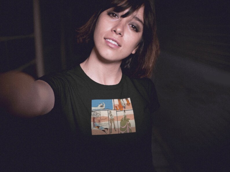 May include: A woman wearing a black t-shirt with a comic book style graphic print of a scene with four panels. The graphic features a person in a white shirt and blue jeans, a person in a red shirt and blue jeans, a person in a green shirt and blue jeans, and a person in a white shirt and blue jeans.