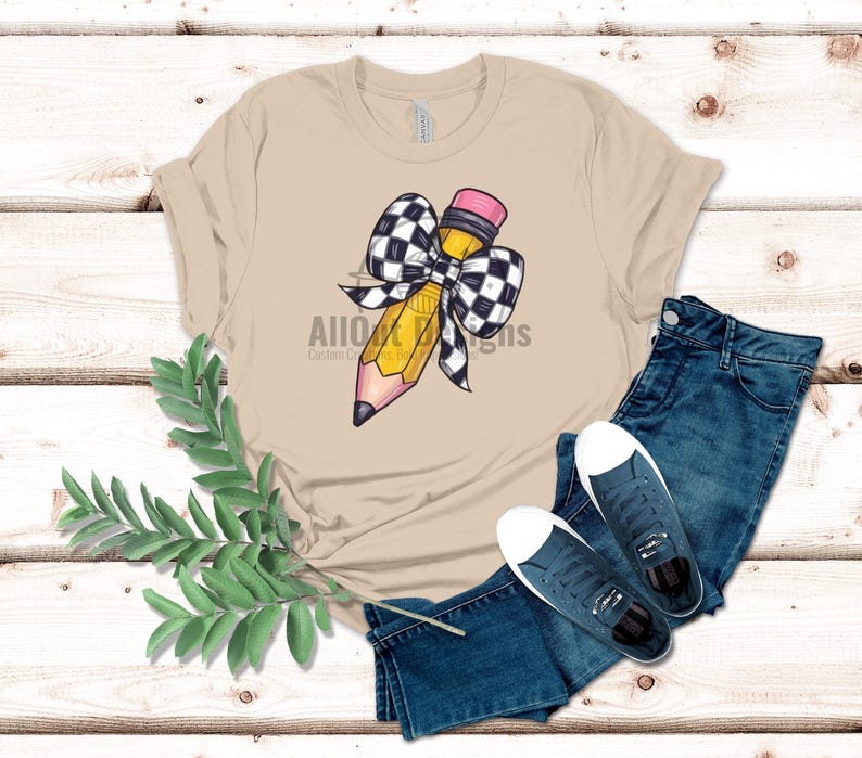 May include: A tan t-shirt featuring a yellow pencil tied with a black and white chequered bow. The pencil has a pink eraser.