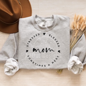 Może przedstawiać: Szara bluza z czarnym kołem z napisem "Stressed Blessed Mom Sometimes A Mess" z sercami wokół koła.