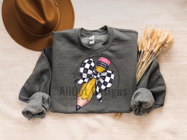 May include: A grey sweatshirt featuring a yellow pencil tied with a black and white chequered bow. The pencil has a pink eraser.
