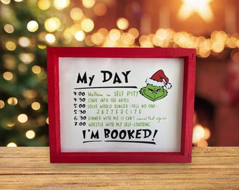 Puede incluir: Un letrero enmarcado en rojo con fondo blanco y un personaje verde del Grinch. El letrero dice "My Day" y enumera una agenda diaria humorística. El letrero dice "I'm Booked!"