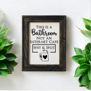 Op de afbeelding: Een ingelijst bord met de tekst "This is a Bathroom Not an Internet Cafe Shit & Split" en een zwart-witte illustratie van een toiletrol met een hart erop.