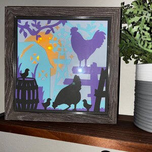 Può includere: Un'opera d'arte intagliata su carta incorniciata che raffigura un gallo viola, una gallina nera e tre pulcini in un ambiente da cortile. La scena è illuminata da una luce gialla calda, creando un'atmosfera fantasiosa e affascinante.