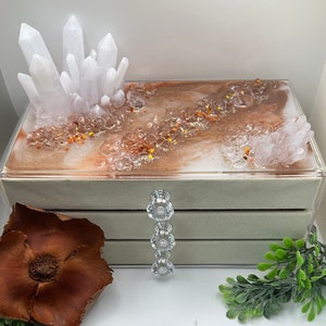 Peut inclure: Une boîte à bijoux à trois niveaux avec un couvercle transparent. Le niveau supérieur présente un motif en résine avec des tourbillons blancs et bruns, des cristaux blancs et des paillettes orange. La boîte a une tirette en forme de cristal.