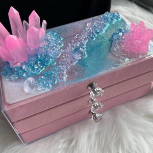 May include: A pink jewelry box with a clear lid, decorated with pink crystal formations and blue and clear decorative elements. The box has three drawers and a clear crystal knob.