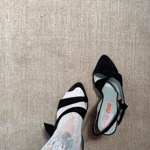 May include: A pair of black pointed-toe slingback heels with a bow detail on the side. One shoe is worn with a white lace stocking, while the other sits separately. The interior sole features the red text Miu Miu and the words Made in Italy.