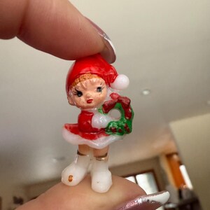 May include: A miniature Christmas figurine of a girl wearing a red dress and hat with white trim, holding a green wreath with a red bow. The figurine has a painted face and white boots. A festive holiday decoration.