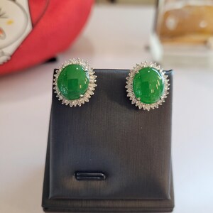 May include: A pair of stud earrings featuring large, round, vibrant green gemstones surrounded by a halo of smaller, clear stones. The earrings are displayed on a black jewelry stand, showcasing their elegant design.