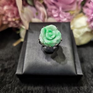 May include: A light green jade ring shaped like a rose. The ring is set on a silver band and sits on a black display box. The background features blurred pink and purple flowers.