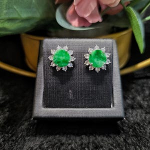 Op de afbeelding: Stud oorbellen met een heldergroene centrale steen, omringd door heldere, sprankelende stenen. De oorbellen zijn gezet in een zilverkleurig metaal. Ze worden gepresenteerd op een zwarte, getextureerde ondergrond, die hun elegante ontwerp benadrukt.