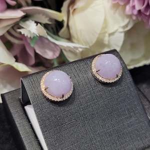 May include: A pair of round lavender-colored stud earrings. Each earring features a light purple, polished stone surrounded by a gold-colored, diamond-studded halo. The earrings are displayed on a black, textured surface, with a blurred floral background.