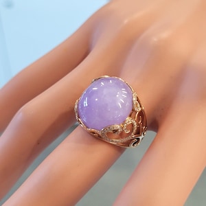 May include: A gold ring featuring a large, oval-shaped, lavender-colored gemstone. The ring has an ornate gold setting with delicate filigree and small, sparkling accent stones. The gemstone is smooth and polished.