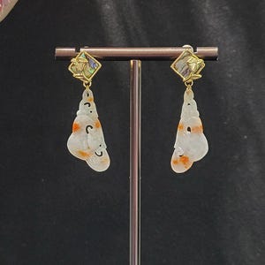 May include: A pair of dangle earrings with a white jade design and a gold-toned abalone shell top. The earrings are displayed on a black stand.