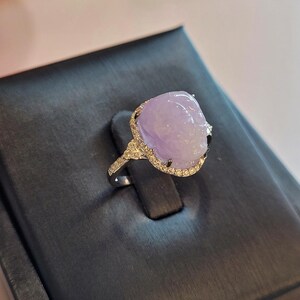 May include: A silver ring featuring a large, oval-shaped, lavender-colored gemstone. The gemstone is surrounded by a halo of small, clear stones. The ring is set on a black display stand.