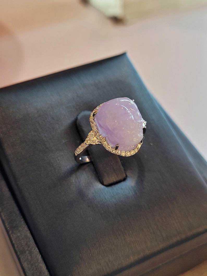 May include: A silver ring featuring a large, oval-shaped, lavender-colored gemstone. The gemstone is surrounded by a halo of small, clear stones. The ring is set on a black display stand.
