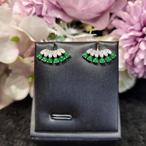 May include: A pair of stud earrings with a fan-shaped design. Each earring features a row of small, round, bright green gemstones at the base, topped with a row of smaller, clear gemstones. The earrings are displayed on a black jewelry stand.