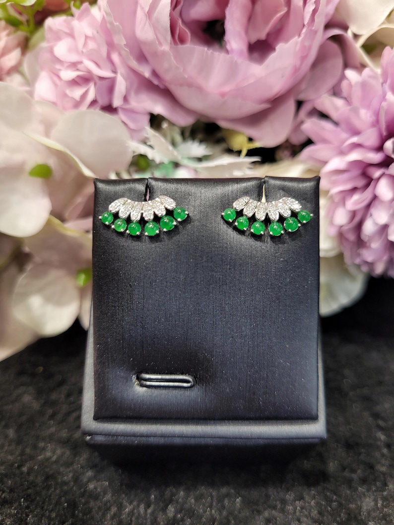 May include: A pair of stud earrings with a fan-shaped design. Each earring features a row of small, round, bright green gemstones at the base, topped with a row of smaller, clear gemstones. The earrings are displayed on a black jewelry stand.