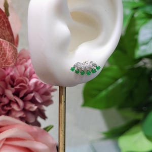 May include: A silver stud earring with a fan-shaped design. The earring features small, round, bright green gemstones and clear, sparkling stones. The earring is displayed on a white ear-shaped form, with a gold-colored stand.