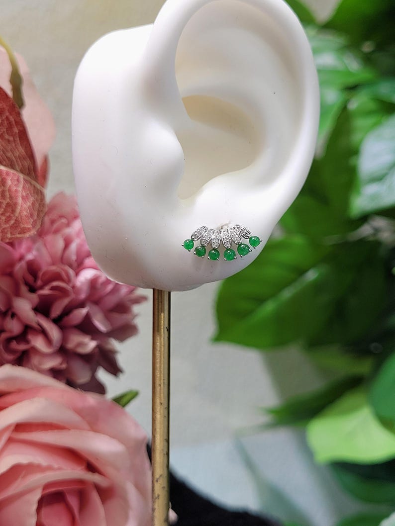 May include: A silver stud earring with a fan-shaped design. The earring features small, round, bright green gemstones and clear, sparkling stones. The earring is displayed on a white ear-shaped form, with a gold-colored stand.