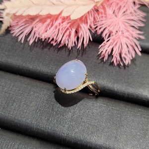 May include: A lavender jade ring with a diamond-encrusted band. The ring is set on a black velvet surface.