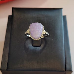 May include: A silver ring featuring a large, oval-shaped, lavender-colored gemstone. The gemstone is surrounded by a halo of small, clear gemstones. The ring is displayed on a black jewelry box.
