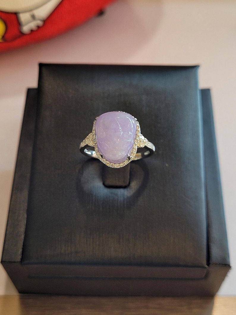 May include: A silver ring featuring a large, oval-shaped, lavender-colored gemstone. The gemstone is surrounded by a halo of small, clear gemstones. The ring is displayed on a black jewelry box.
