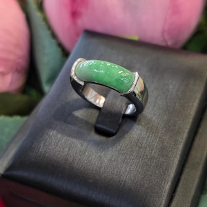 May include: A ring featuring a smooth, polished, cylindrical green jade stone set horizontally into a polished silver-toned metal band. The band has a solid, thick construction with a reflective finish that curves around the finger.