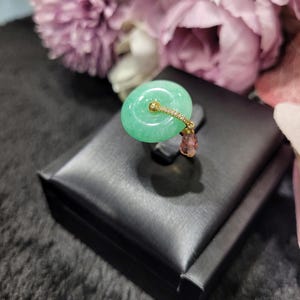 May include: A jade pendant with a light green circular jade stone, a gold-colored band with small clear stones, and a small pinkish-brown stone. The pendant is displayed on a black box with a blurred floral background.