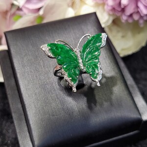 May include: A butterfly ring with a vibrant green jade butterfly design. The ring features a silver band and is accented with small, sparkling gemstones along the wings. The ring is displayed on a black surface with a blurred floral background.