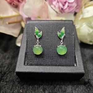 May include: A pair of dangle earrings featuring a round, light green gemstone. The earrings have a silver-colored metal design with green leaf accents at the top. The earrings are displayed in a black jewelry box.